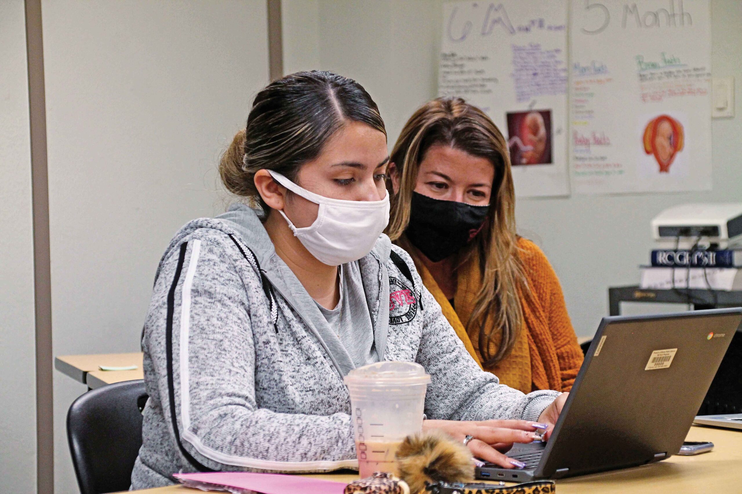 Two women with masks using a laptop in classroom.