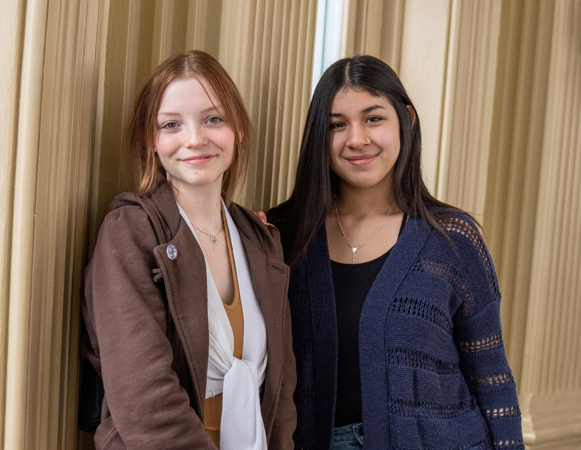 Two women smiling indoors, leaning against a wall.